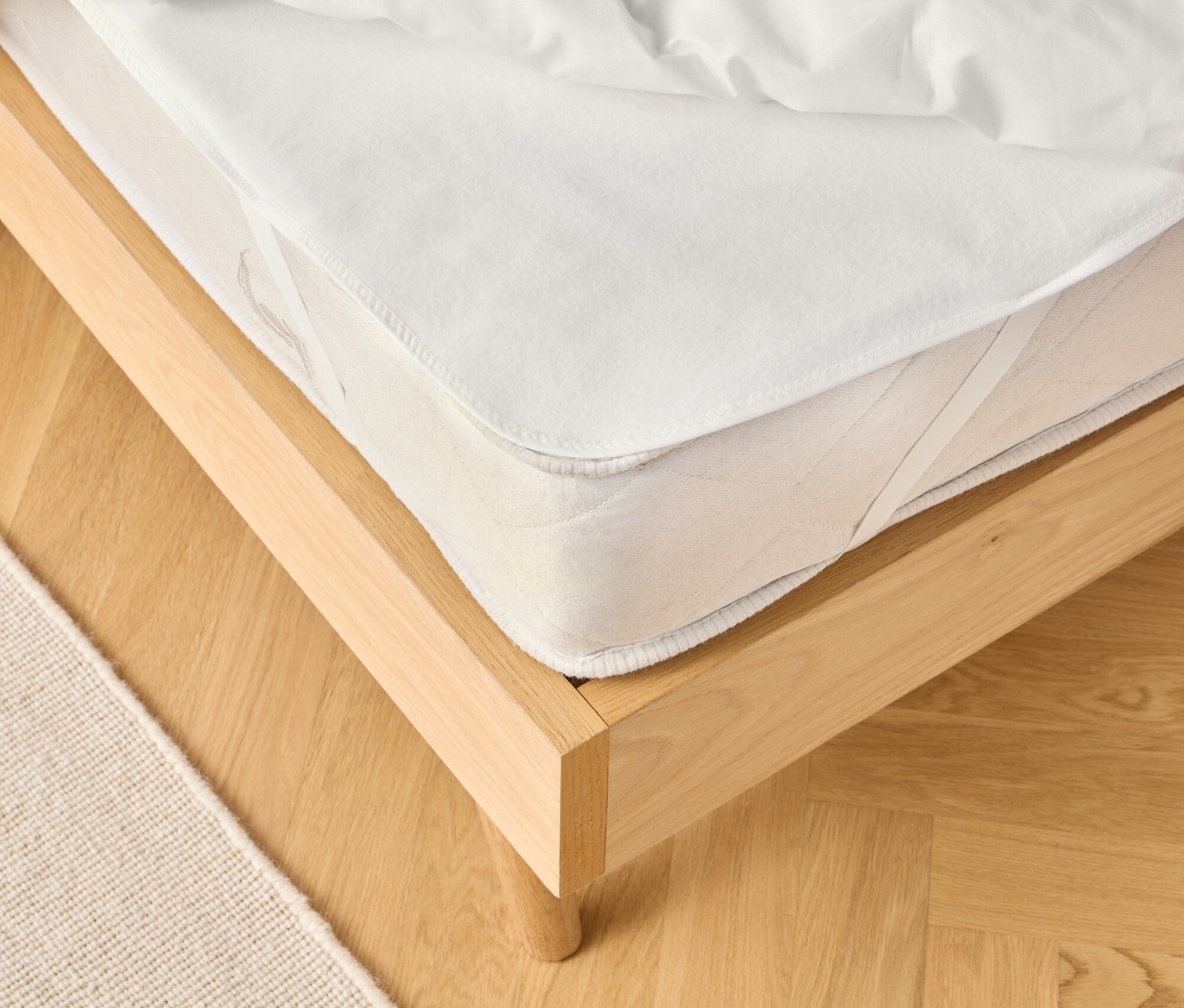 Kilátás egy világos fa ágykeretre fehér matraccal és fehér paplannal. Egy bézs szőnyeg részben látható.