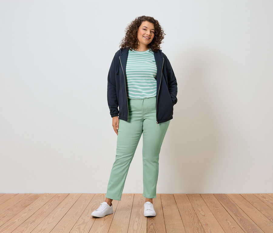 Egy göndör hajú nő áll a fal előtt, sötétkék kabátot, csíkos pólót, zöld nadrágot és fehér cipőt visel.