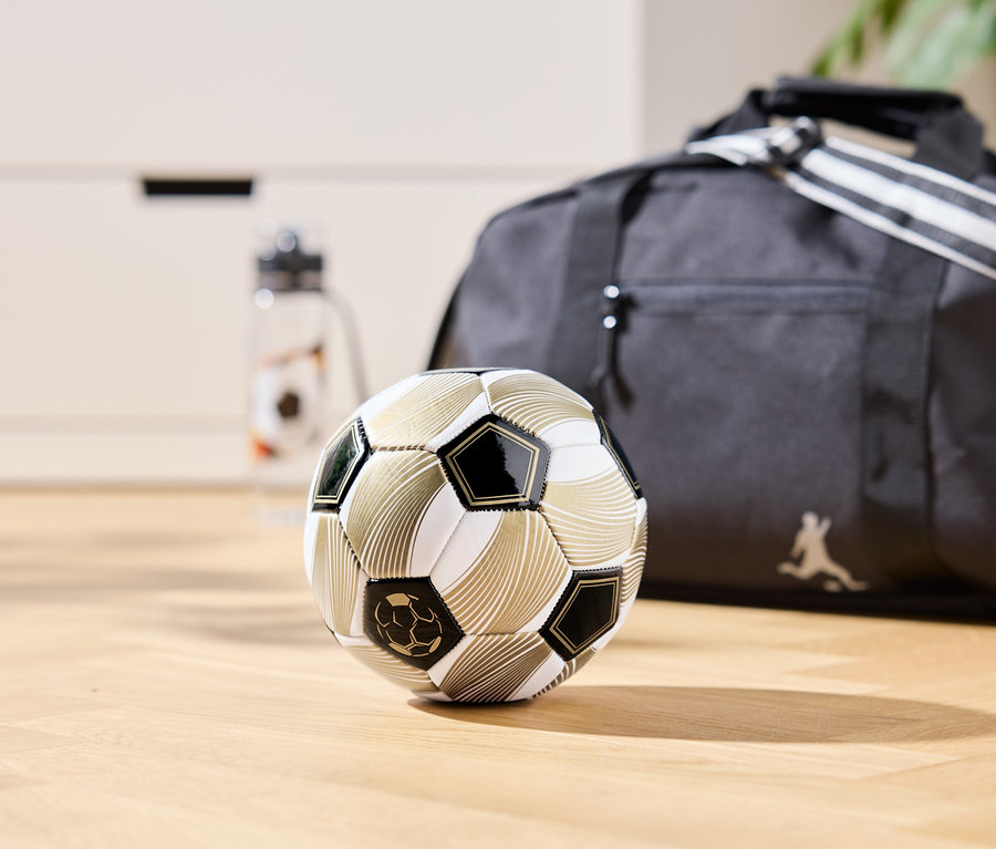 Egy arany színű focilabda fekszik egy fa padlón, a háttérben egy sporttáska és egy vizes palack látható.