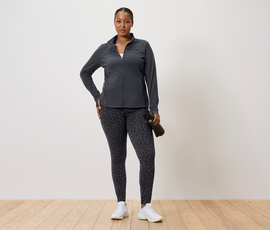 Nő sötét edződzsekiben és leopárdmintás leggingsben, vizet tartalmazó palackot tart.