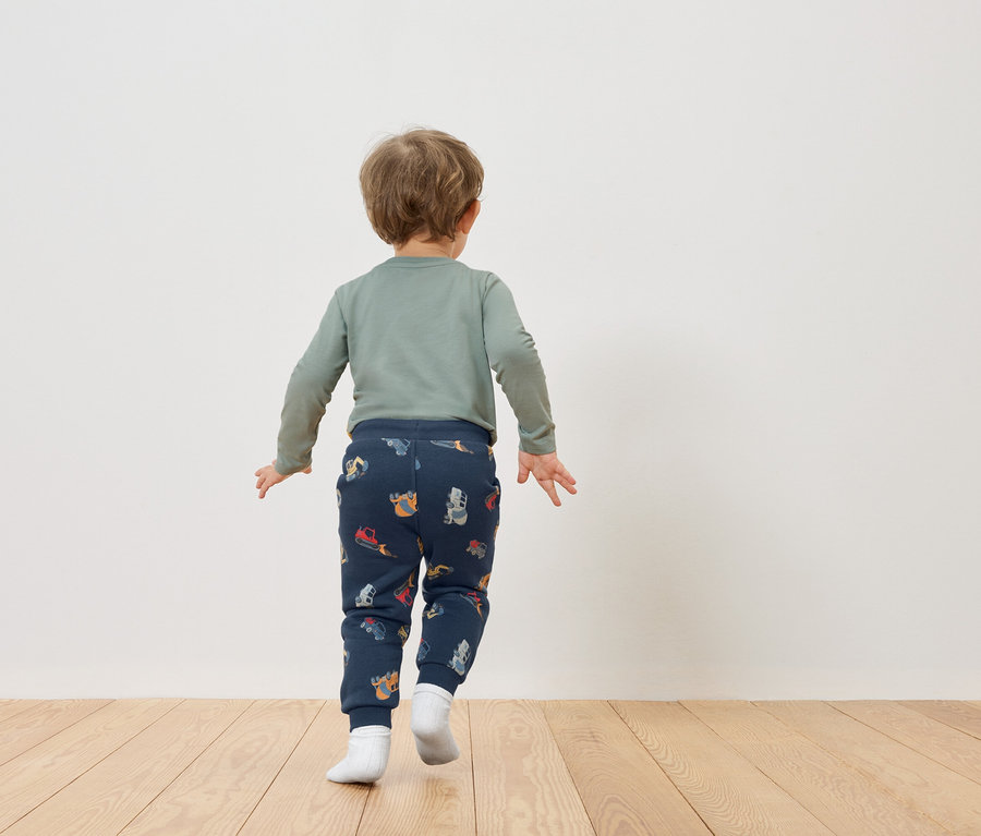 Egy szőke hajú kisgyermek hátulról látható, zöld hosszú ujjú inget és sötétkék, járműmintás nadrágot visel.