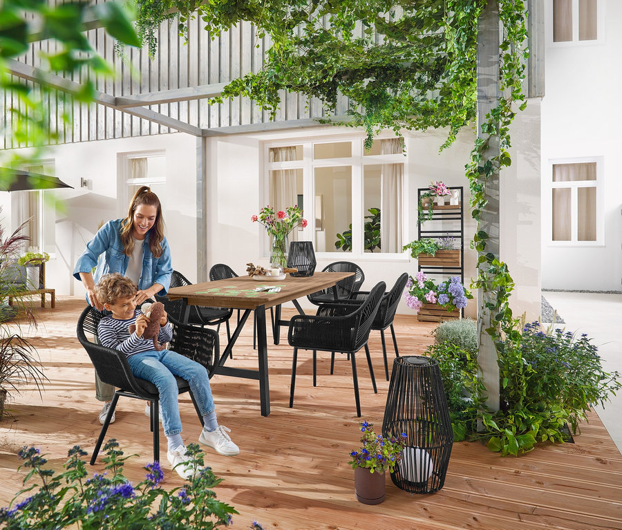 Anya és gyermeke egy kb. 200–250 cm-es, rusztikus kerettel ellátott kihúzható kerti asztalnál ülnek. Egy kicsi és egy nagy Napelemes viharlámpa textilszalaggal áll a földön. A háttérben egy »Elin« virágládás fal látható, tetszés szerint befüggeszthető polcokkal.