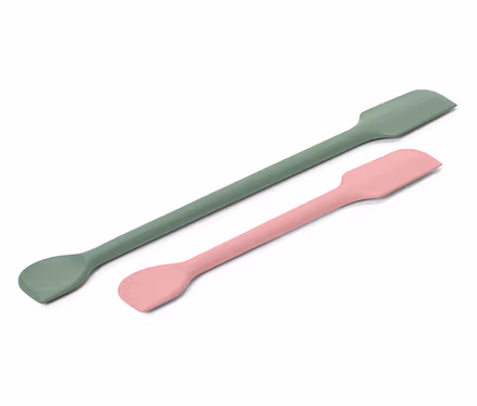 Egy zöld és egy rózsaszín spatula átlósan fekszik egy fehér háttéren.