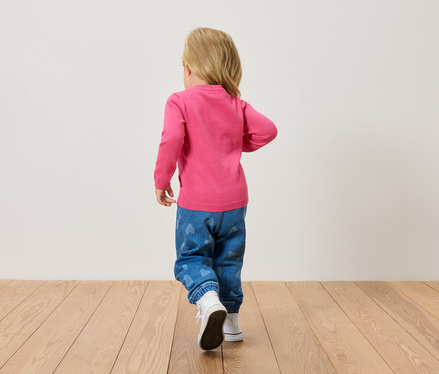 Egy kis szőke gyerek hátulról látható, rózsaszín pulóvert, szívecskés kék farmert és fehér cipőt visel, fa padlón sétál.