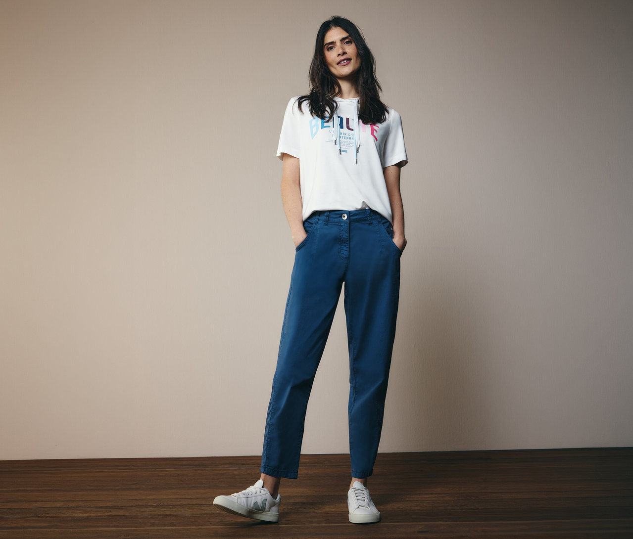 Nő CECIL kapucnis pólóban és kék CECIL »Scarlett« hordónadrágban áll a kamerának, kezét a zsebébe dugva.