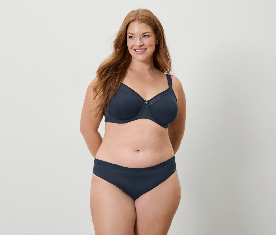 Hosszú vörös hajú nő sötétkék melltartót és bugyit visel, mosolyog.