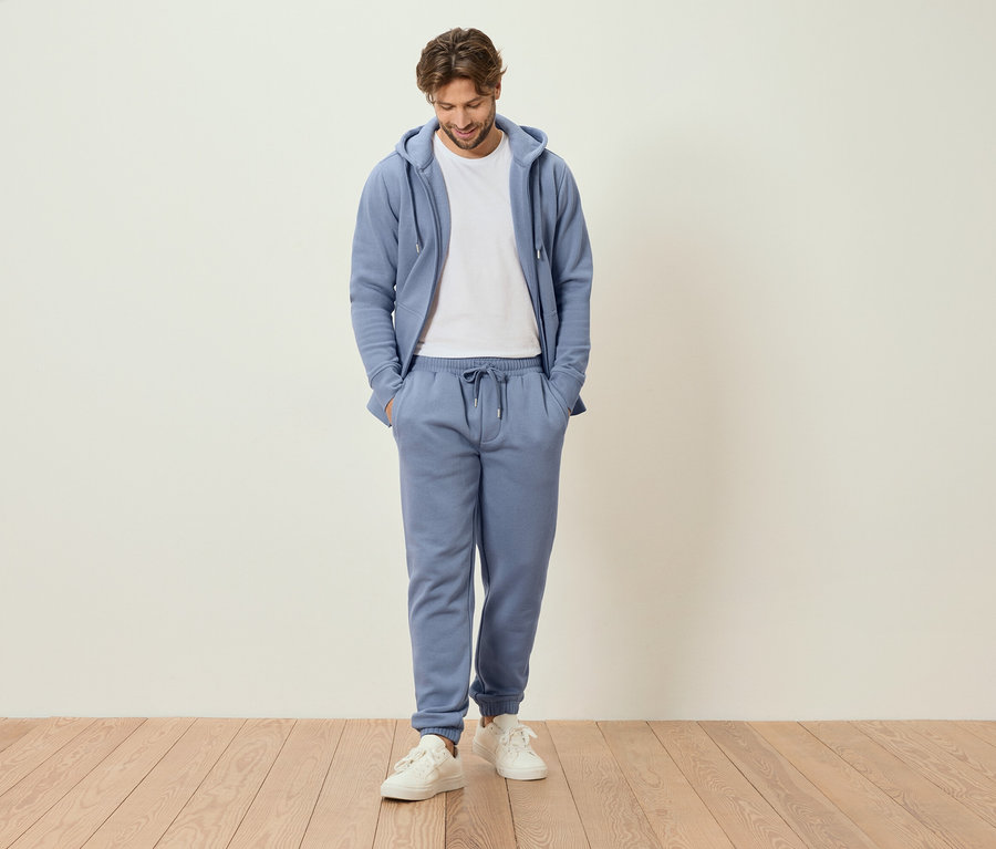 Férfi áll kézzel a zsebében világoskék tréningruhában és fehér tornacipőben.