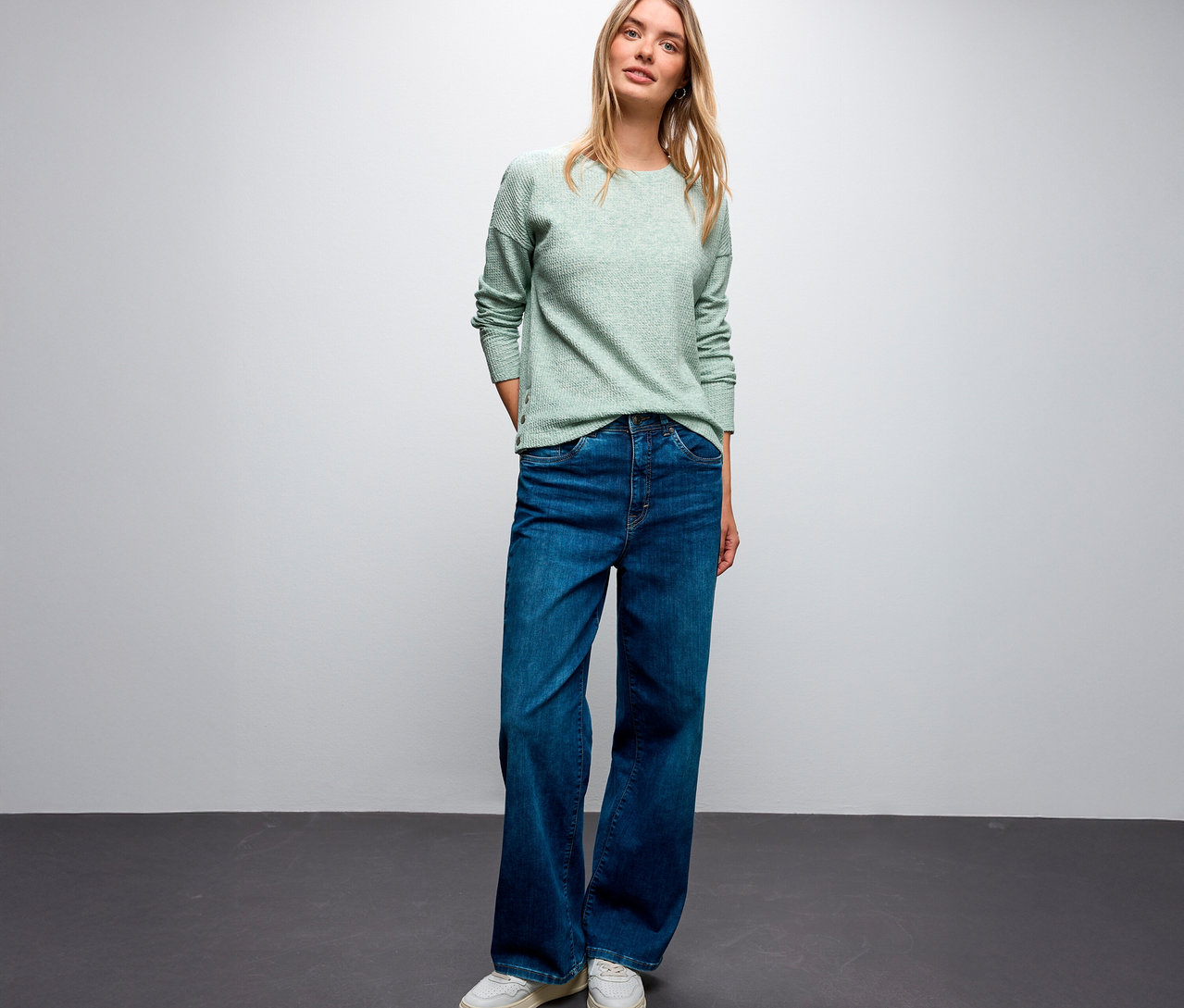 Szőke nőn Street One bordás mintás póló, kék farmer és fehér tornacipő van.