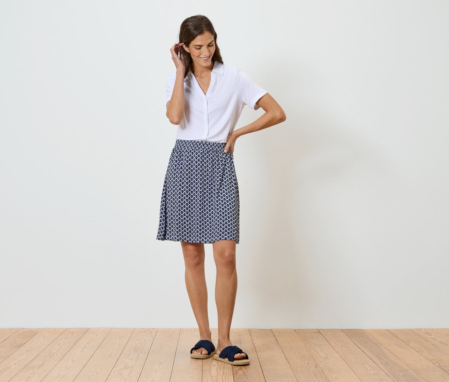 Nő áll egy fehér fal előtt, fehér inget és kék-fehér mintás szoknyát visel.