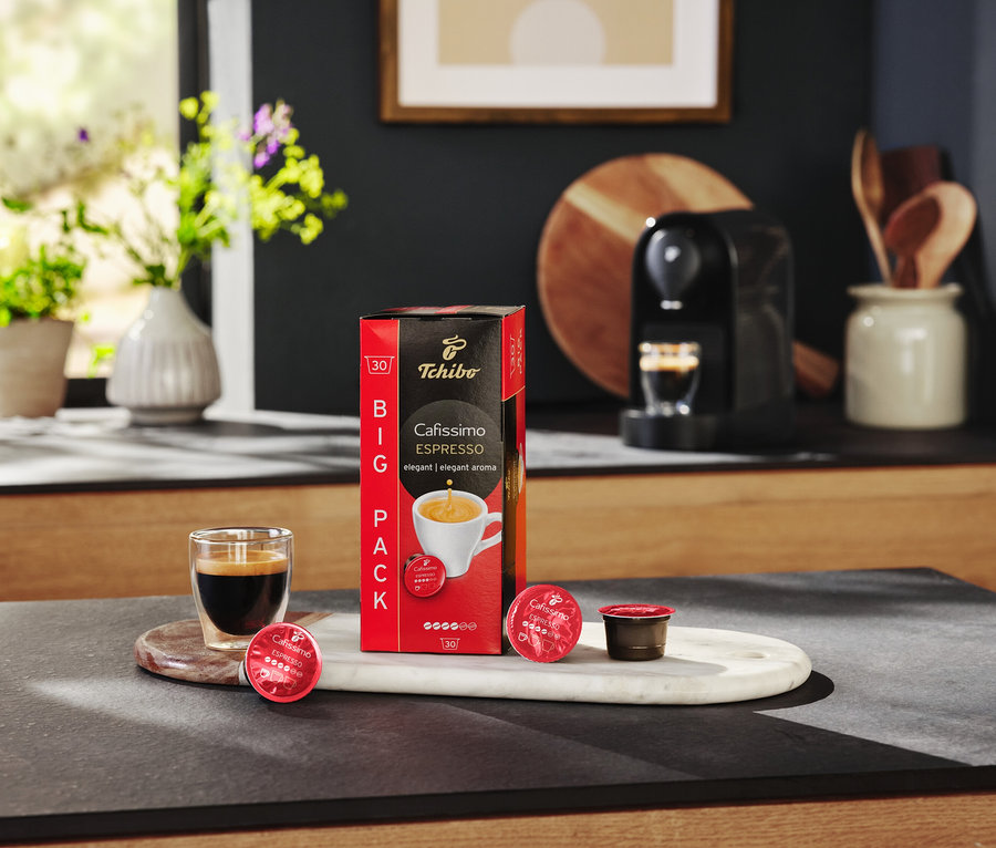 Az asztalon egy csomag Cafissimo Espresso kapszula, egy eszpresszóval teli pohár és egy kávéfőző található.
