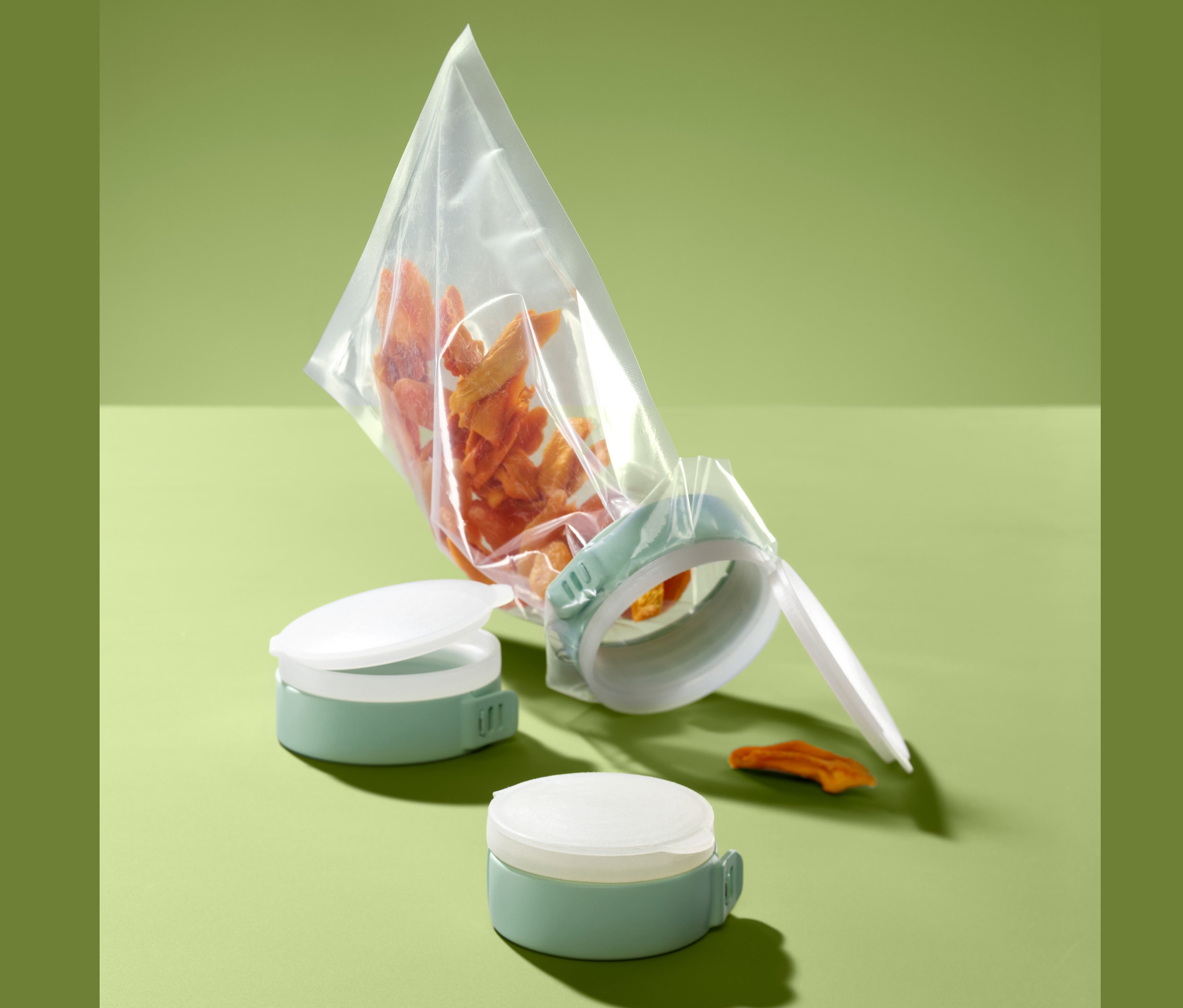 Egy átlátszó zacskó narancssárga ételdarabokkal van lezárva egy zöld 3 tasakzáró gyűrű szettben segítségével. Két további gyűrű látható mellette.