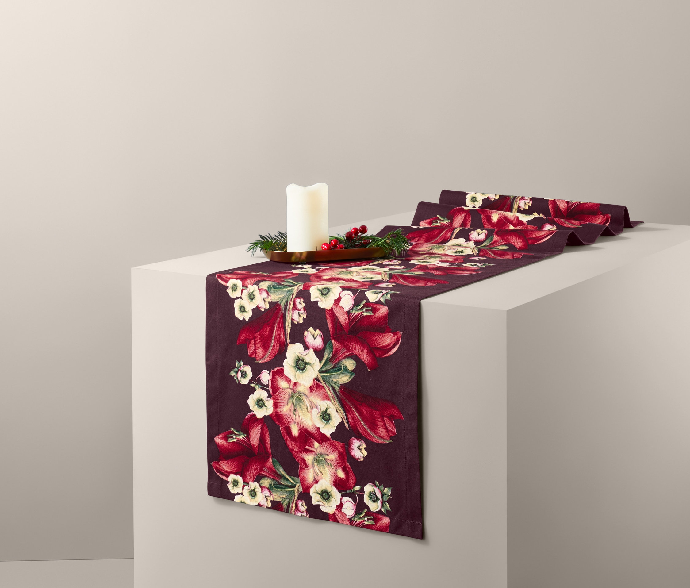 Egy fehér talapzaton bordó, virágos asztali futó és egy gyertya látható.