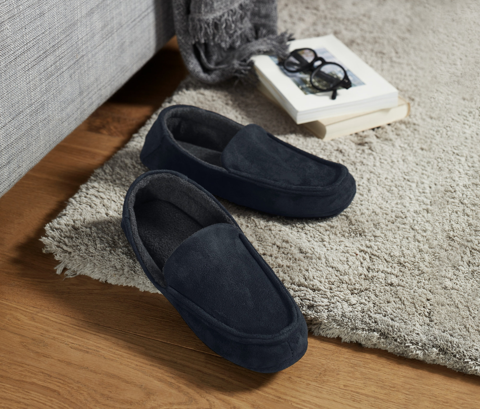 Egy pár sötétkék, velúr hatású férfi házicipő egy bézs szőnyegen, egy kanapé mellett.