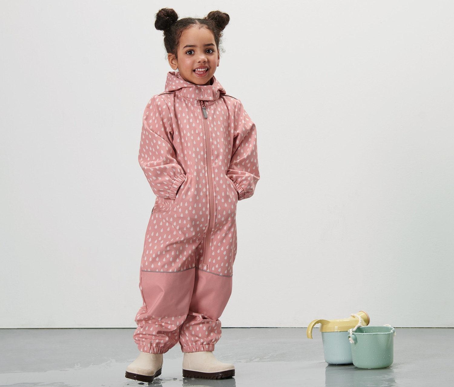 Egy kislány áll egy rózsaszín cseppes, kisgyerek softshell kezeslábasban.