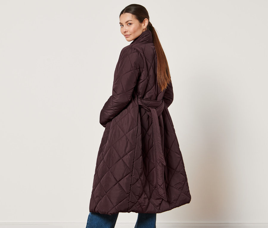 Egy nő a válla fölött hátra néz, és egy női steppelt kabátot visel övvel, padlizsán színben.