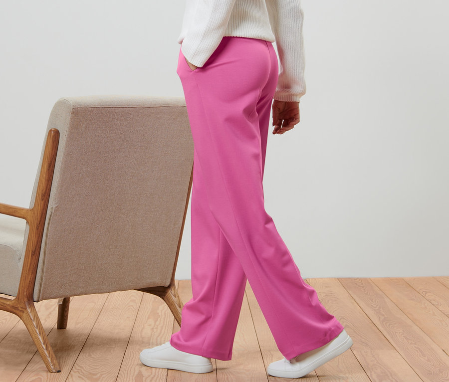 Egy személy rózsaszín, levarrt hajtású jersey nadrágban sétál egy bézs fotel mellett.