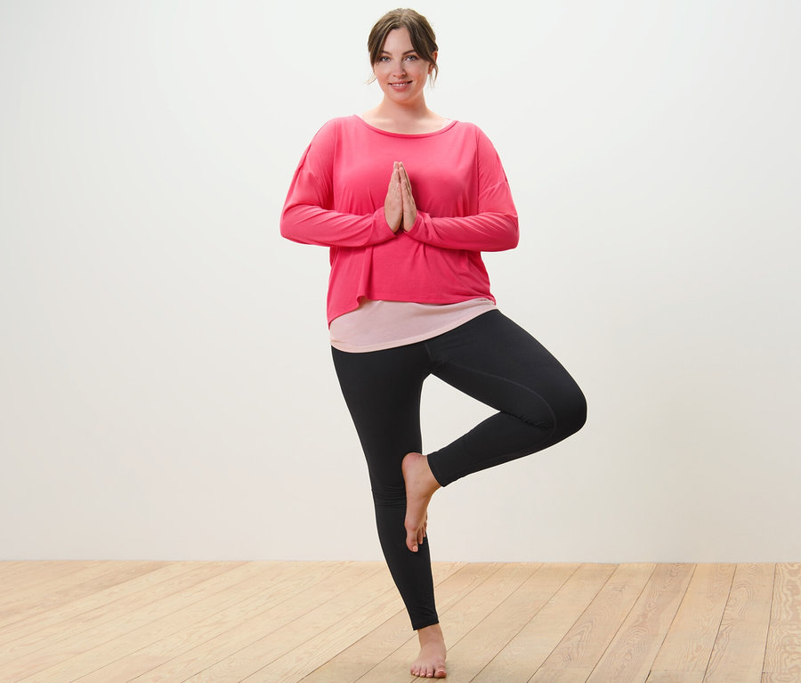 Nő rózsaszín 2 az 1-ben sportfelsőben és fekete sportnadrágban fa pózban áll.