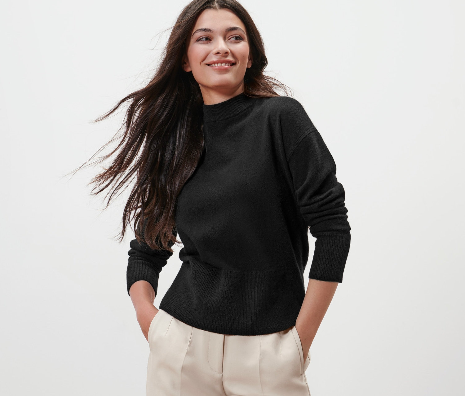 Hosszú barna hajú nő fekete kötött pulóverben (gyapjú/kasmír) és bézs nadrágban pózol.