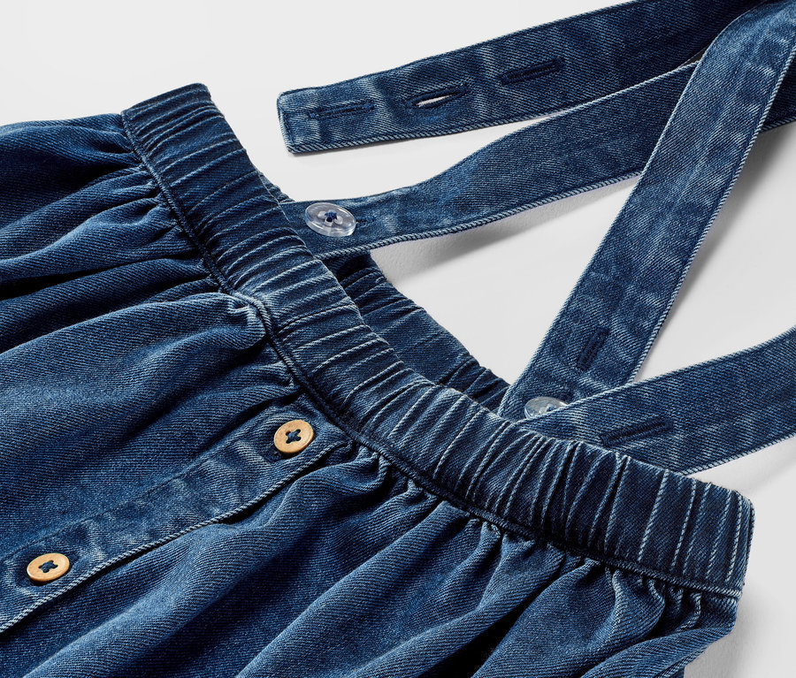 Közelkép egy kisgyerek farmer szoknyáról, pántokkal és gombokkal.