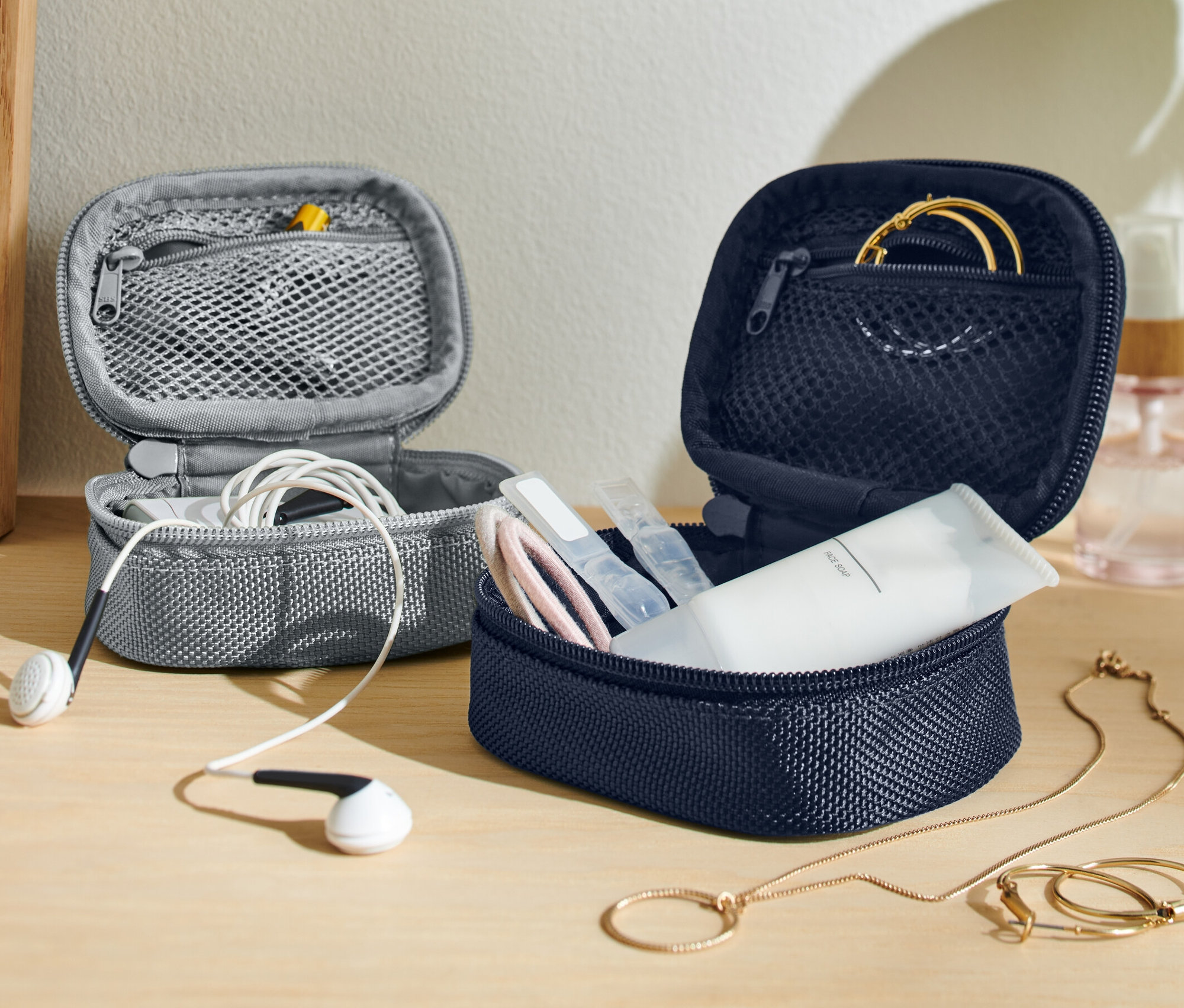Két nyitott 2 mini rendszerezőtáska utazáshoz, egy szürke fehér fülhallgatóval és egy sötétkék kozmetikumokkal és arany ékszerekkel.