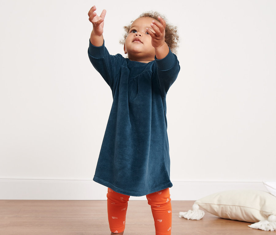 Egy kislány szappanbuborékok után nyúl. Sötétkék plüssruhát és narancssárga leggingset visel.