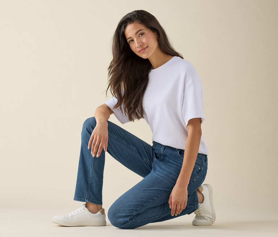 Egy nő féltérden állva mosolyog, fehér pólót, kék farmert és fehér tornacipőt visel.