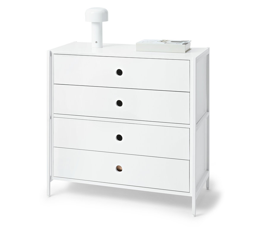 Fehér komód négy fiókkal, a tetején egy lámpa és egy könyv.