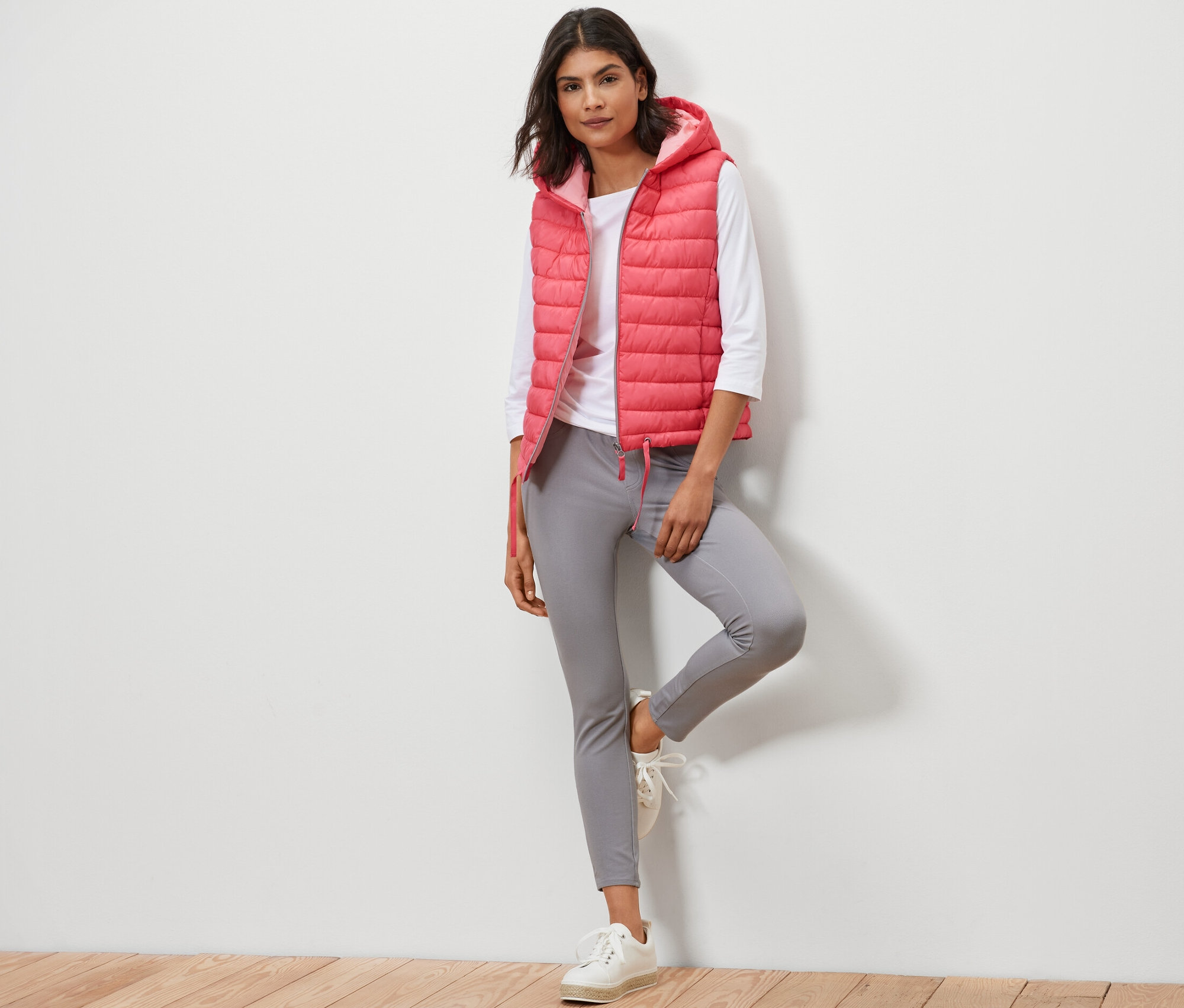 Nő pózol kapucnis steppelt mellényben, fehér pólóban és szürke nadrágban.