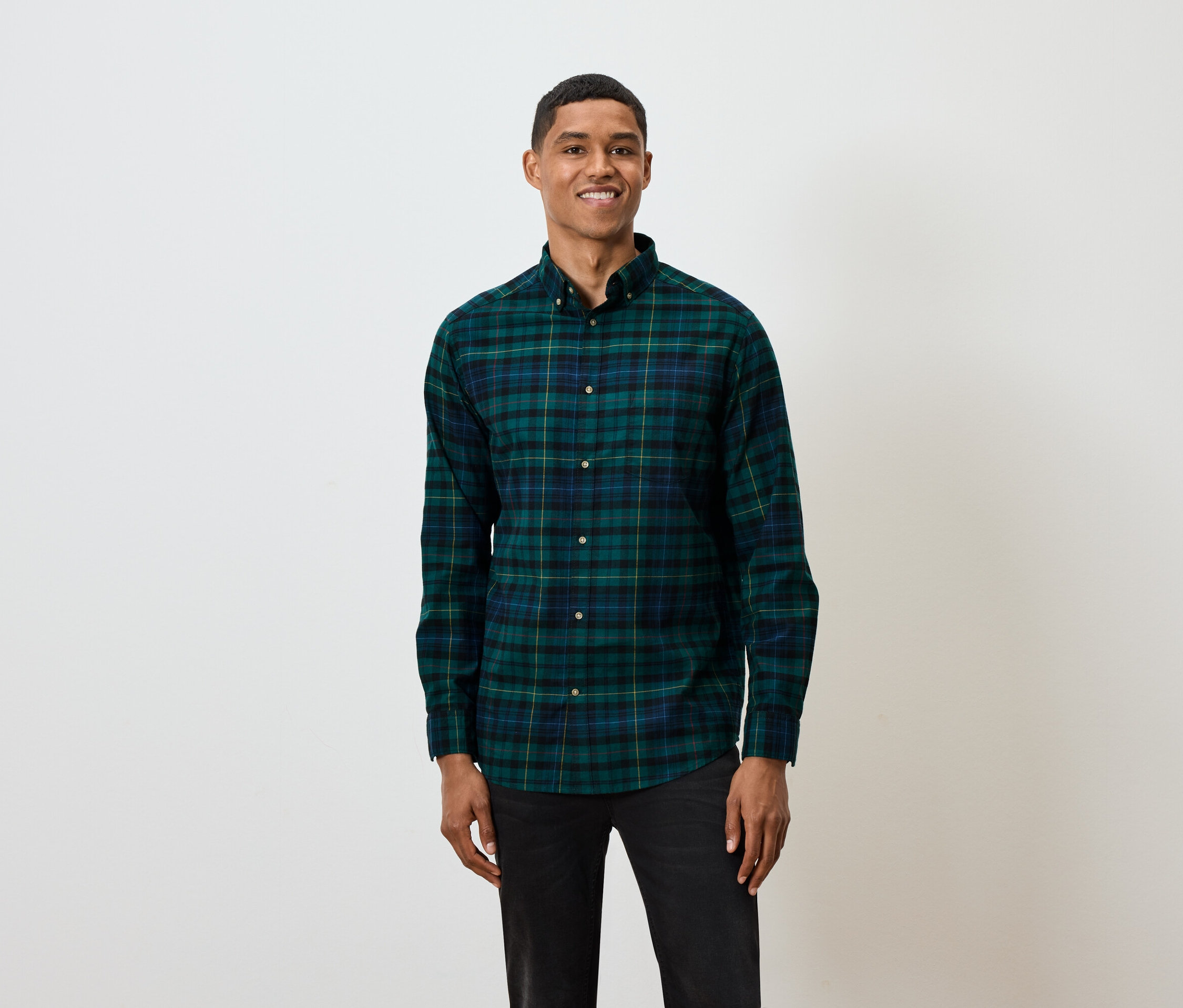 Egy rövid hajú férfi pózol fehér háttér előtt, zöld és kék kockás, hosszú ujjú inget és fekete farmert visel.