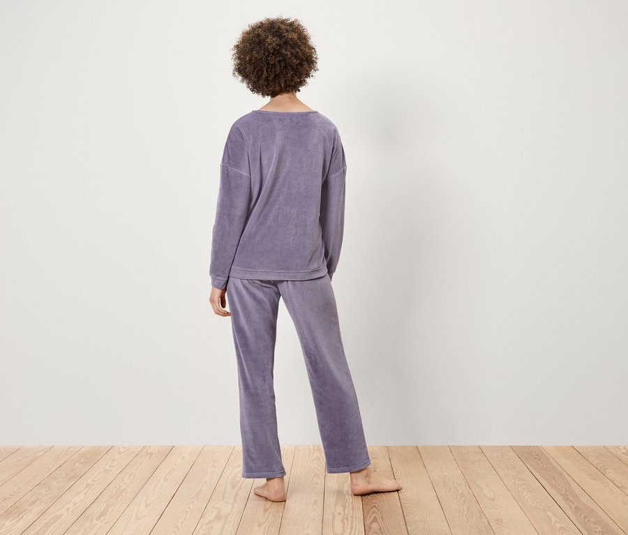 Egy göndör hajú nő áll háttal, lila bársony pizsamát visel világos fa felületen.