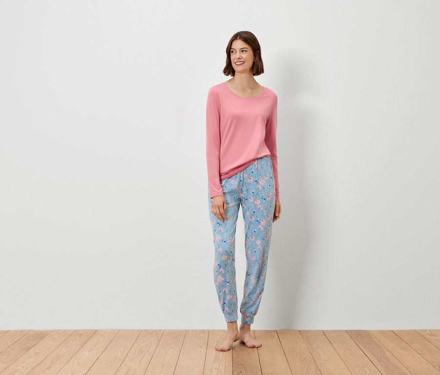 Egy nő áll, rózsaszín hosszú ujjú felsőt és virágmintás kék nadrágot visel.