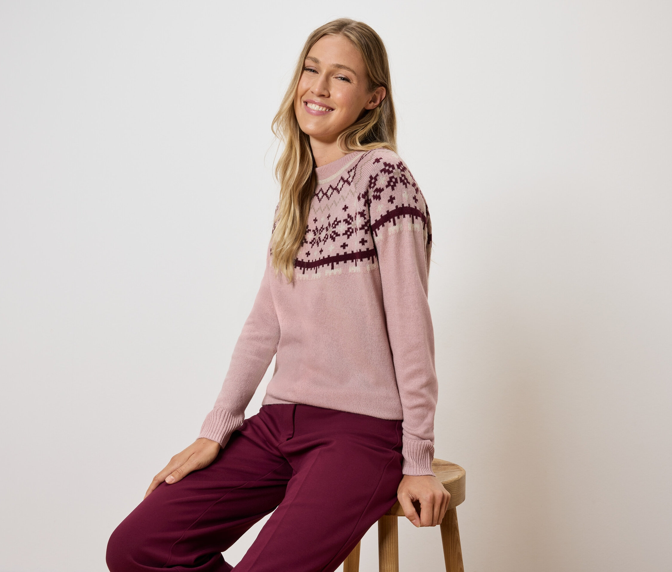 Egy nő ül egy széken, rózsaszín mintás pulóvert és bordó nadrágot visel.