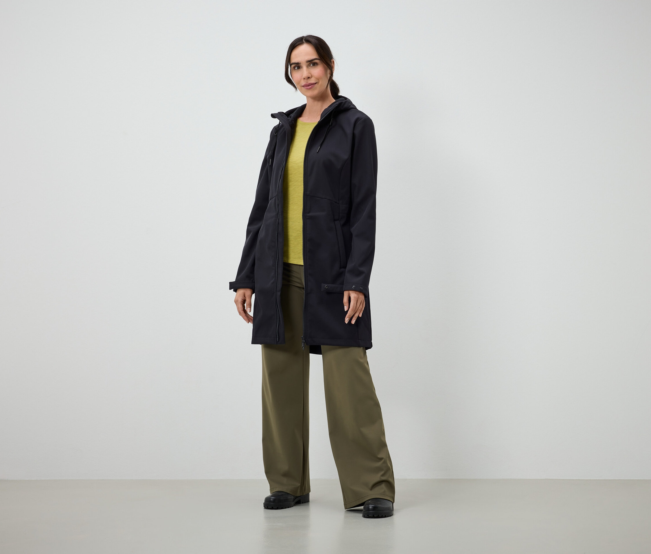 Egy nő áll egy hosszú fekete kabátban, sárga pólóban és bő zöld nadrágban egy beltéri helyszínen.