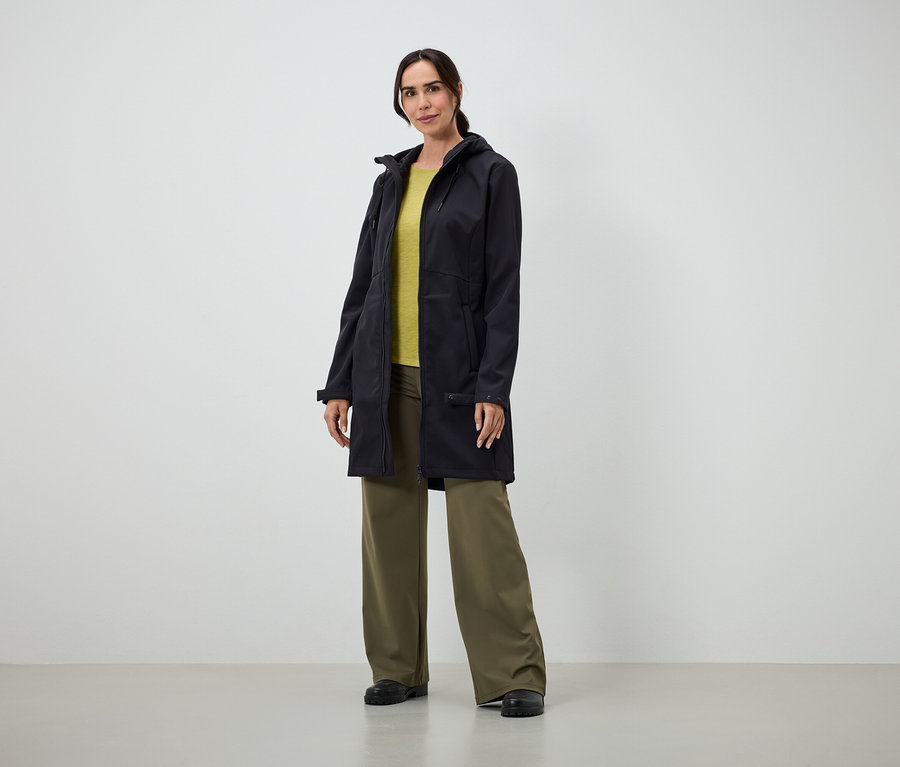 Egy nő áll egy hosszú fekete kabátban, sárga pólóban és bő zöld nadrágban egy beltéri helyszínen.