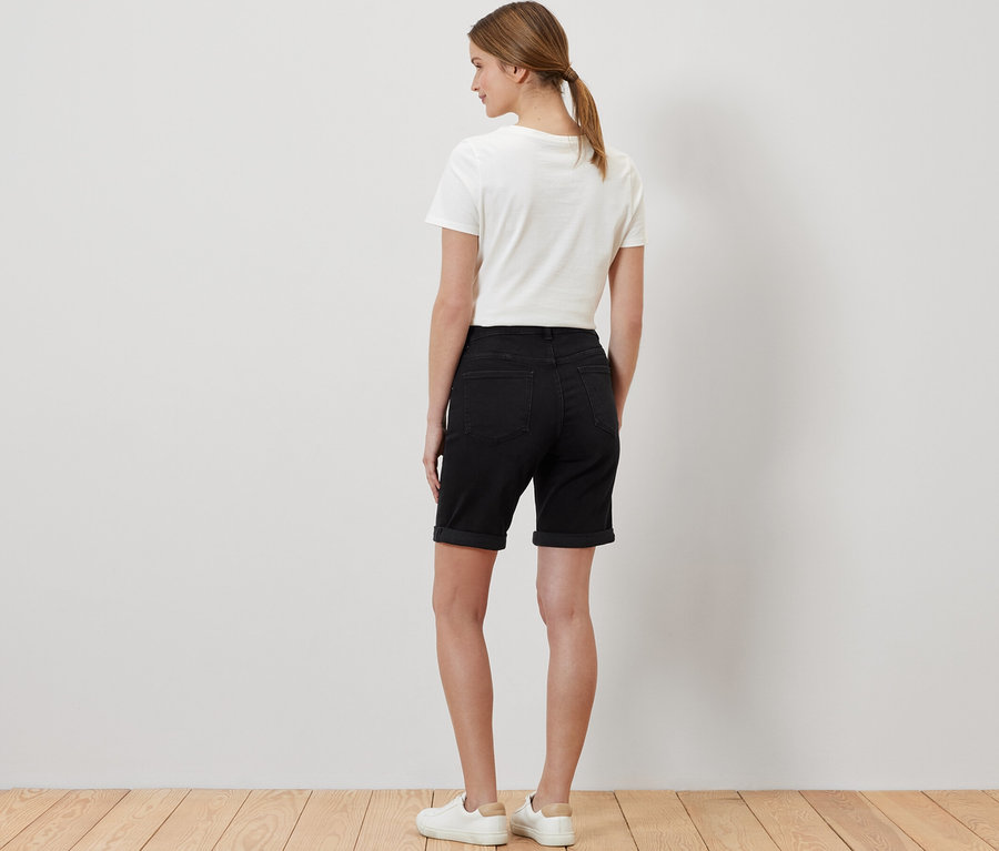 Hátulról látható nő fekete farmer Bermuda »Lea« nadrágban, fehér pólóban és tornacipőben.