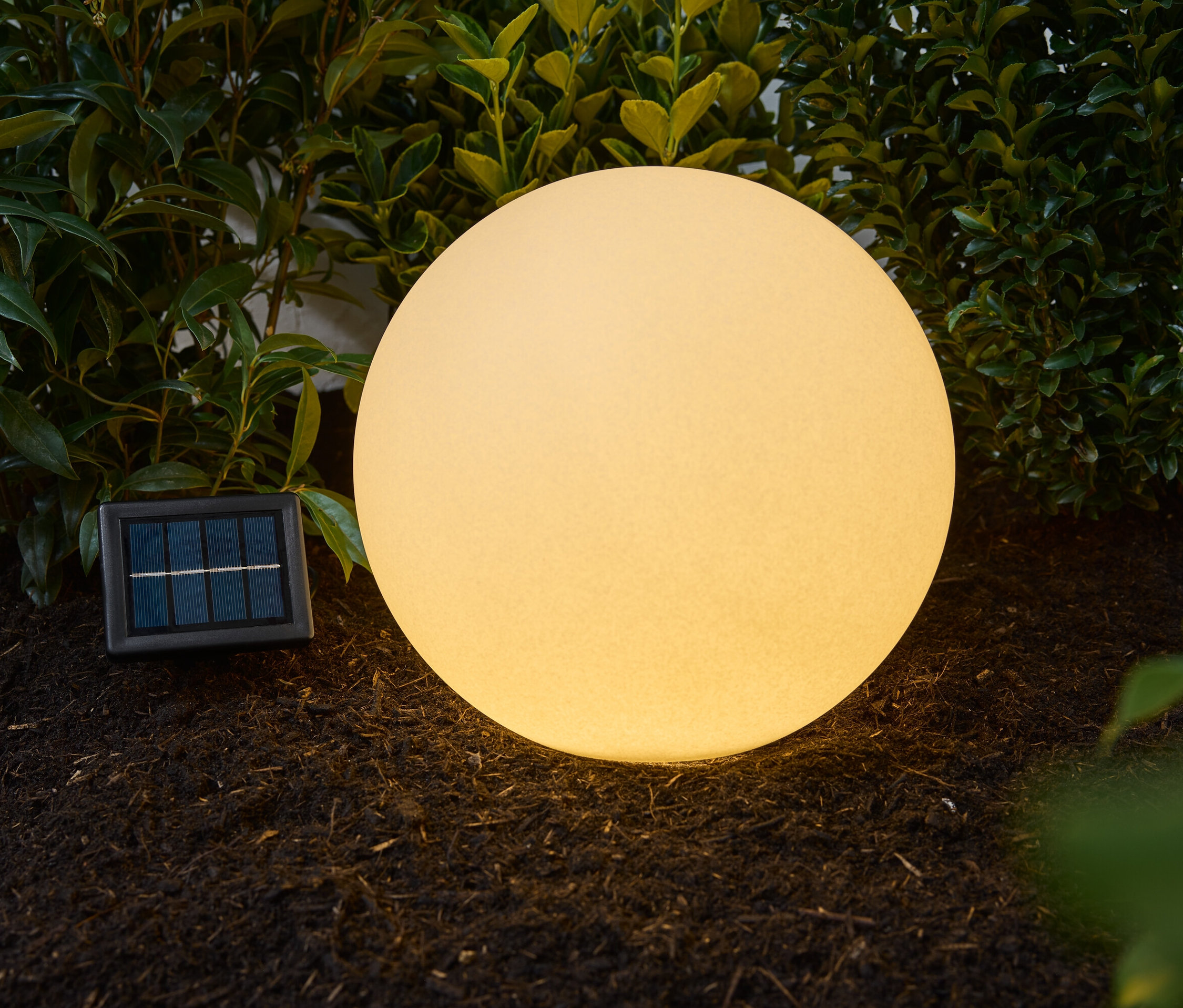 Egy kb. 30 cm-es, homok hatású napelemes gömblámpa áll a szabadban a földön, zöld növények mellett.
