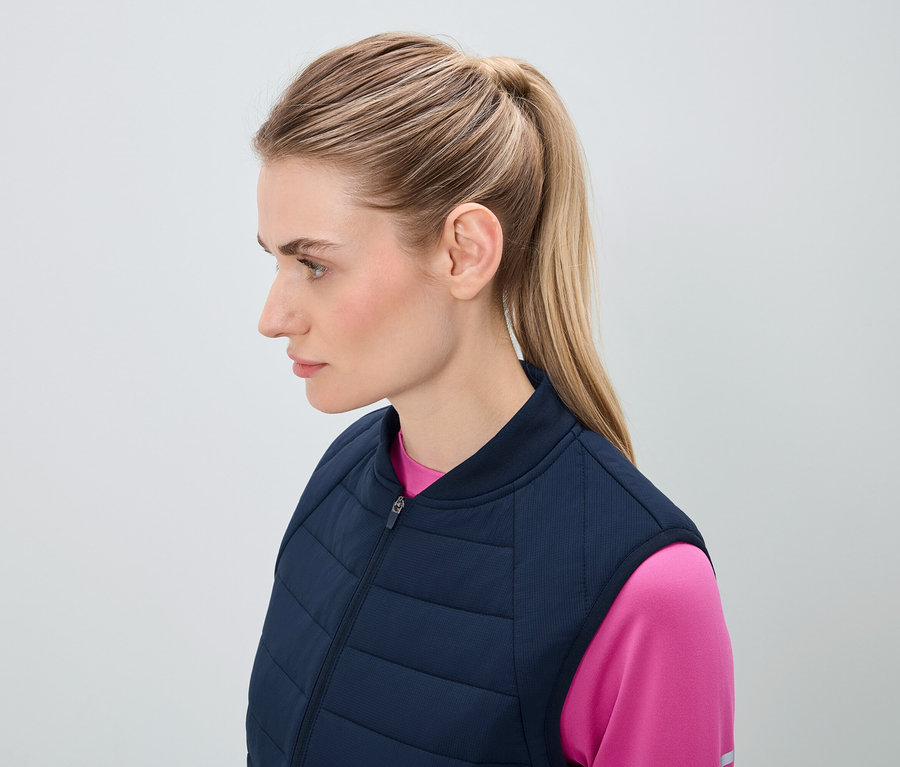 Profil egy szőke hajú, lófarokba kötött nőről, sötétkék steppelt mellényben és rózsaszín felsőben.