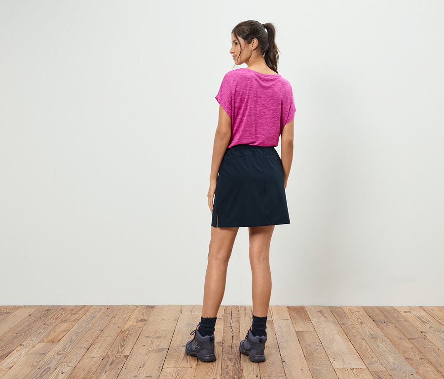 Egy lófarokba kötött hajú nő háttal áll, rózsaszín pólót, fekete szoknyát és szürke cipőt visel.