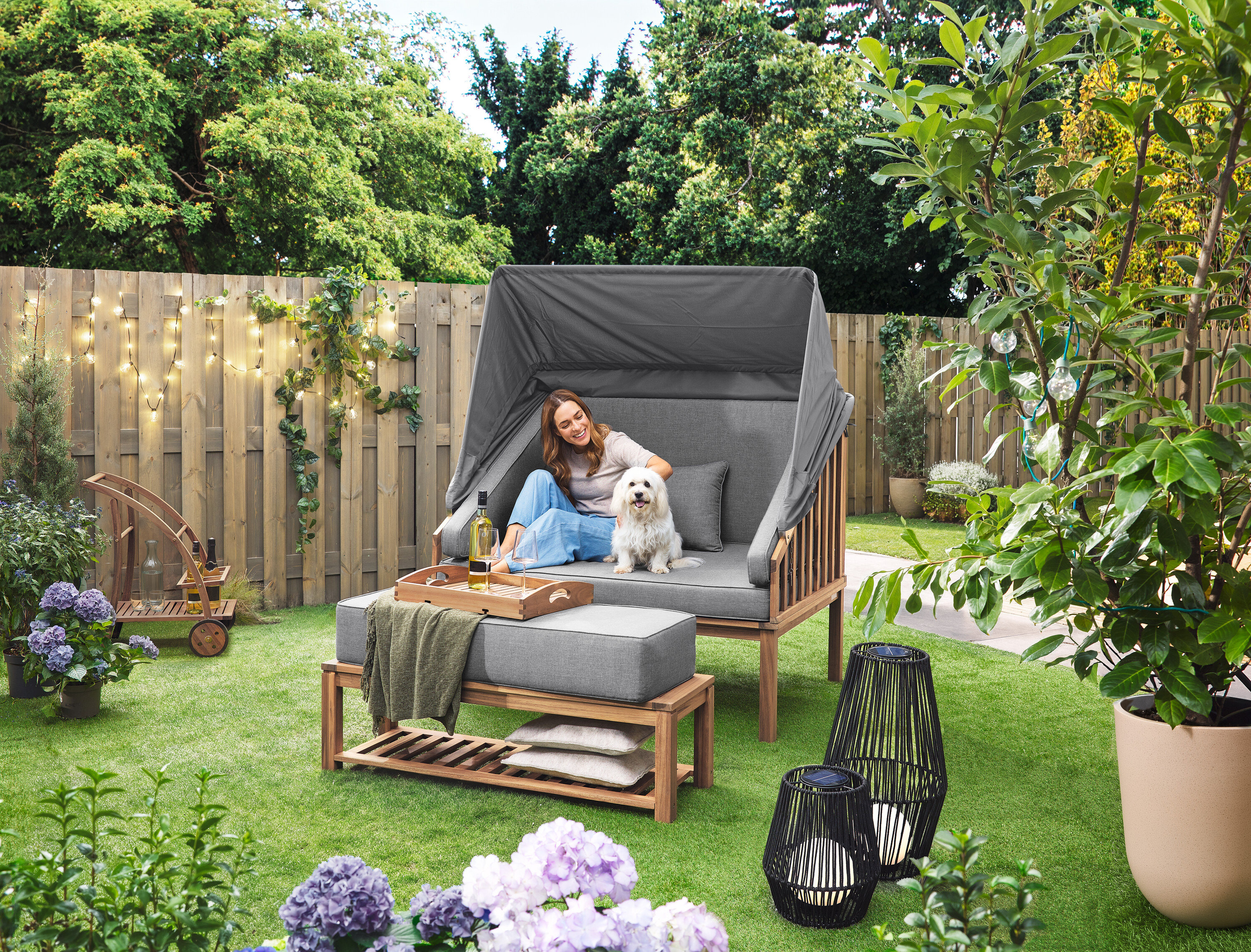 Egy nő ül egy kutyával egy Premium Lounge-Strandkorb mit Sonnenschutz-Dach nevű strandszékben. A füvön egy nagyméretű, textilpántos napelemes viharlámpa és egy kisméretű, textilszalaggal ellátott napelemes viharlámpa áll. A háttérben egy levehető tálcás bárkocsi látható. Egy LED-es kültéri fényfüzér lóg a kerítésen.