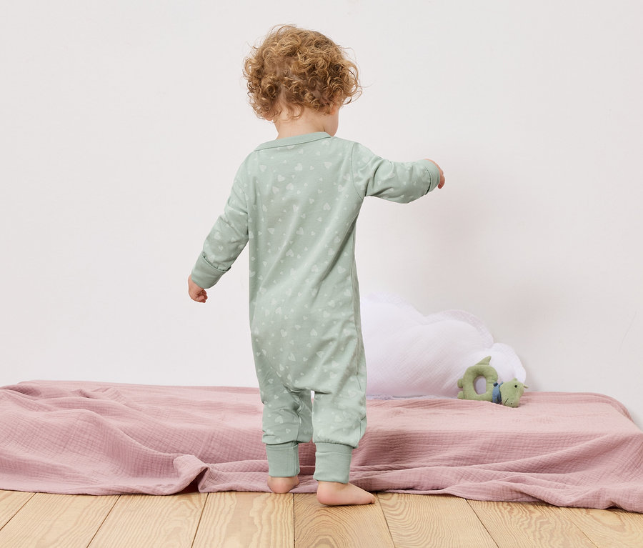 Egy kisgyermek háttal áll a kamerának egy ágyon, rózsaszín takaróval és egy zöld játékkal.