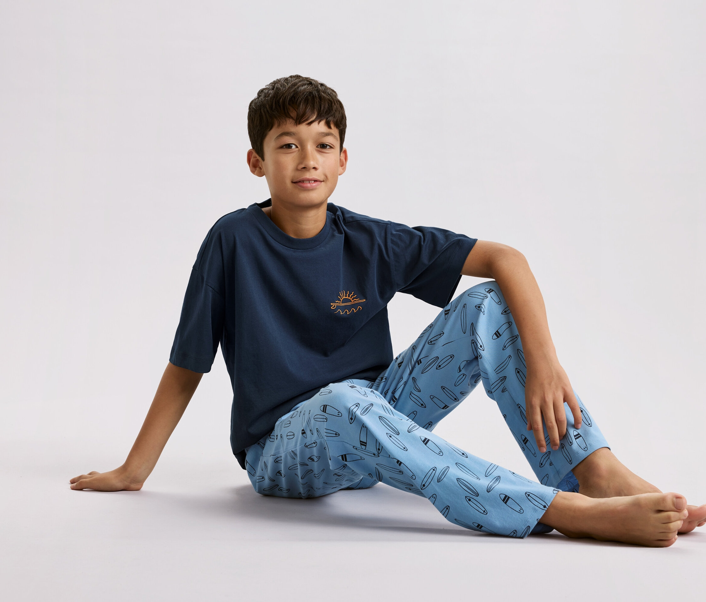 Egy fiú ül a földön, tengerészkék pólót visel, rajta egy kis nap és hullámok, valamint világoskék pizsamanadrágot szörfdeszka mintával.