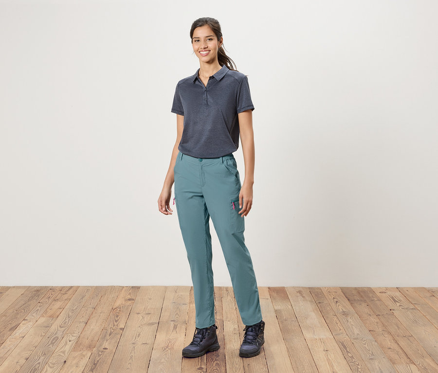 Egy nő áll egy fehér fal előtt, kék pólót, kék nadrágot és túrabakancsot visel.