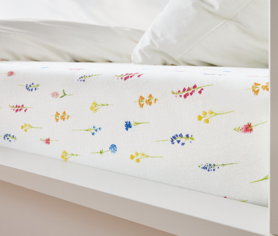 Közelkép egy ágyról, amelyen fehér lepedő van, sárga, kék, narancssárga és rózsaszín virágmintával.