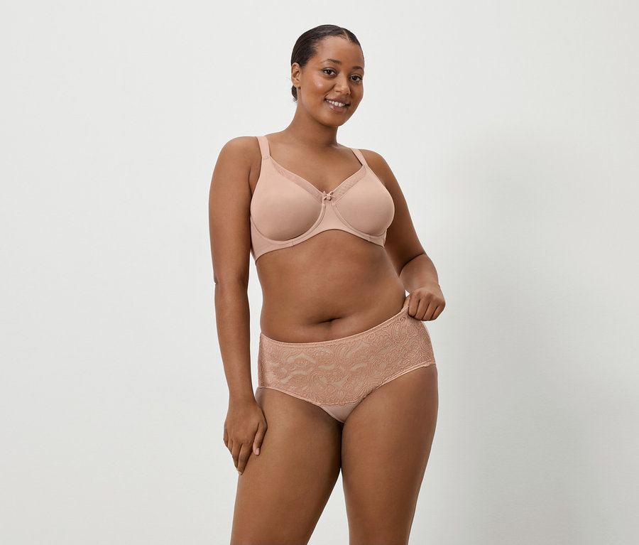 Sötét bőrű nő visel testszínű melltartót és hozzáillő csipkés bugyit egyszerű háttér előtt.