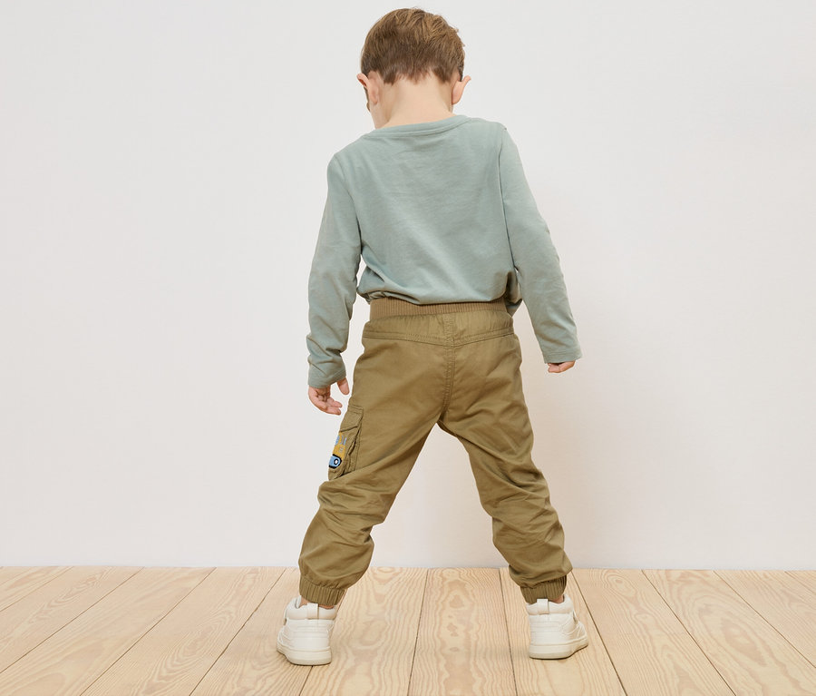 Egy gyermek áll háttal a kamerának, zöld hosszú ujjú pólót, khaki nadrágot és fehér tornacipőt visel.