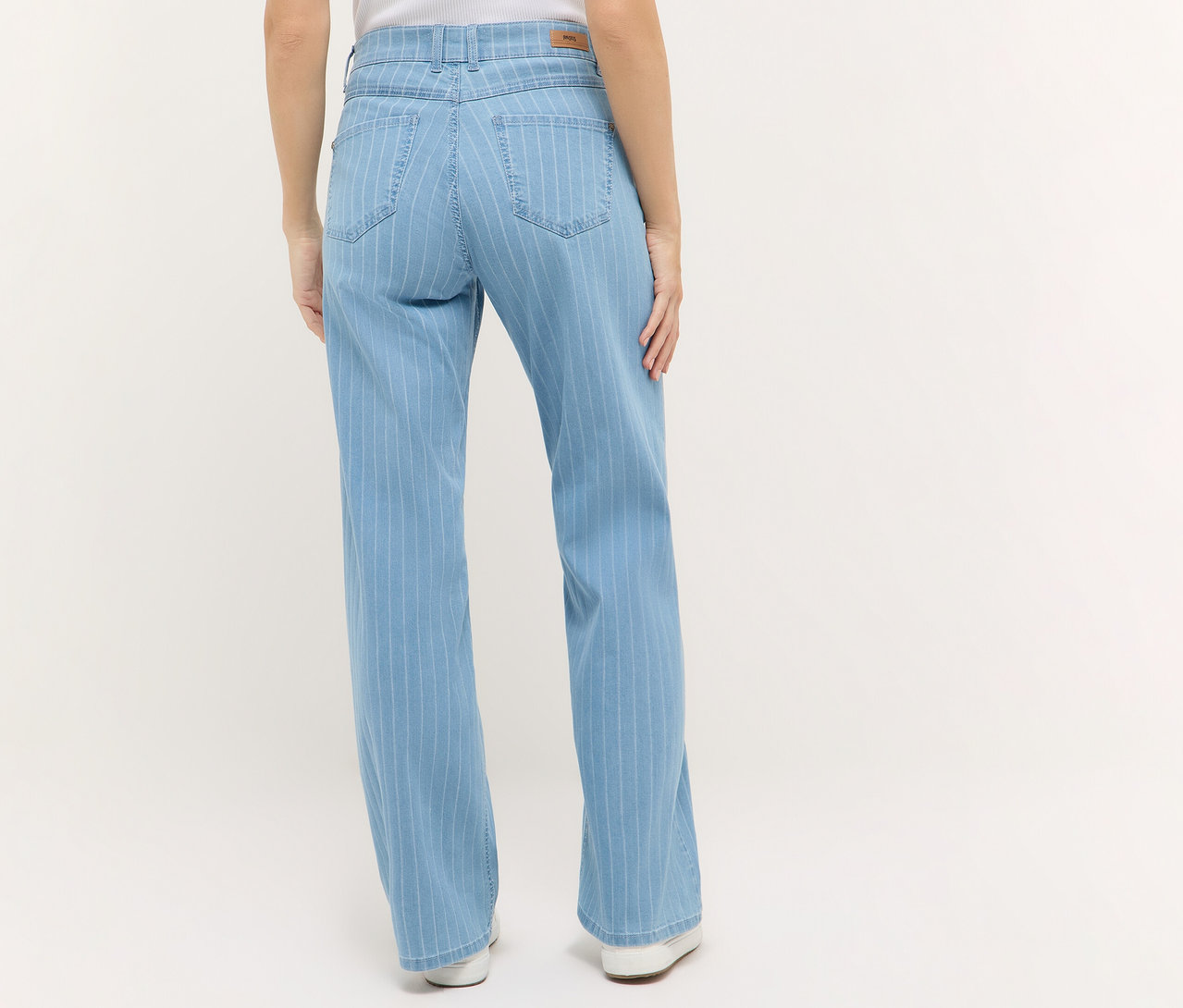 Hátulnézet egy világos csíkos Angels denim »Liz« bőszárú farmernadrágot és fehér cipőt viselő nőről világos háttér előtt.