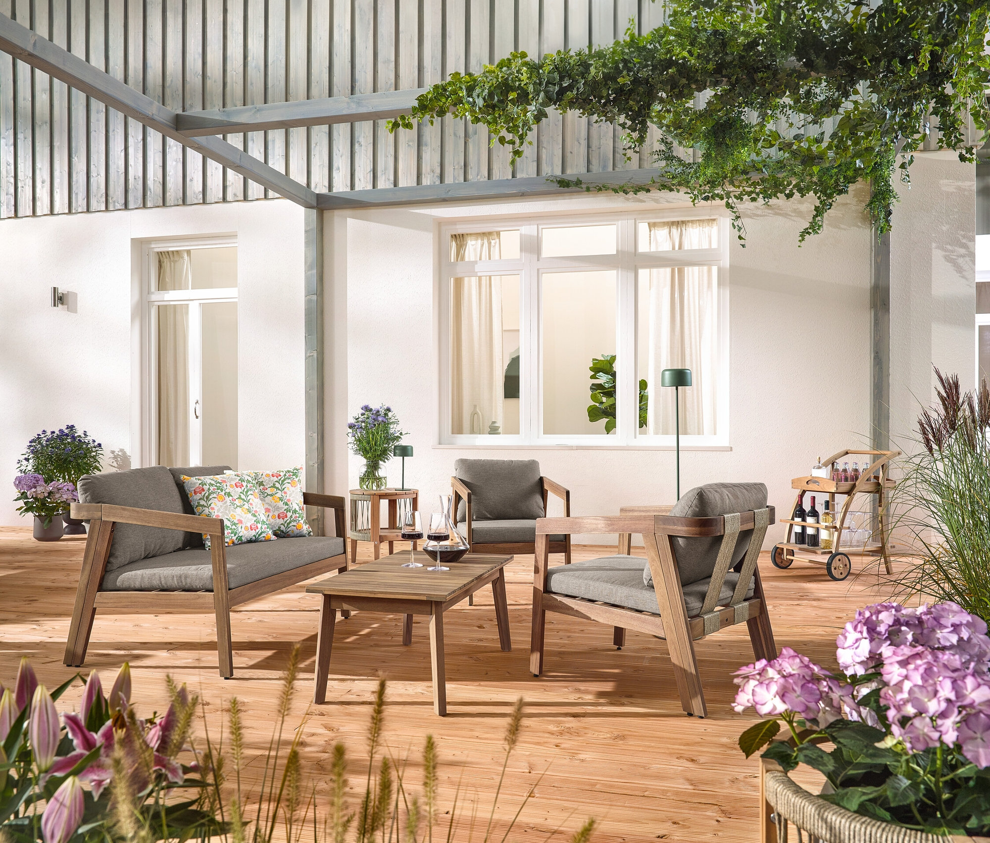 Egy fa teraszon egy 4 részes, textilfonatos kerti ülőgarnitúra áll, 2 virágmintás kültéri párnával, egy bárkocsi levehető tálcával és egy kerti asztal levehető asztallappal, Ø kb. 50 cm. Egy szolár akkus asztali lámpa, dzsungelzöld és egy szolár akkus állólámpa, dzsungelzöld is látható.