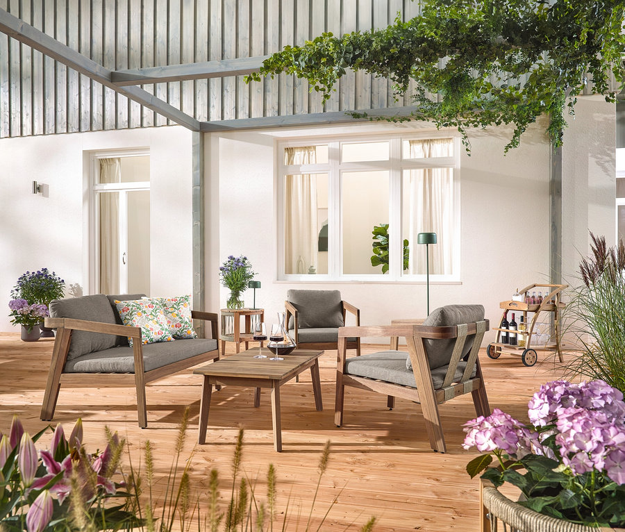 Egy fa teraszon egy 4 részes, textilfonatos kerti ülőgarnitúra áll, 2 virágmintás kültéri párnával, egy bárkocsi levehető tálcával és egy kerti asztal levehető asztallappal, Ø kb. 50 cm. Egy szolár akkus asztali lámpa, dzsungelzöld és egy szolár akkus állólámpa, dzsungelzöld is látható.