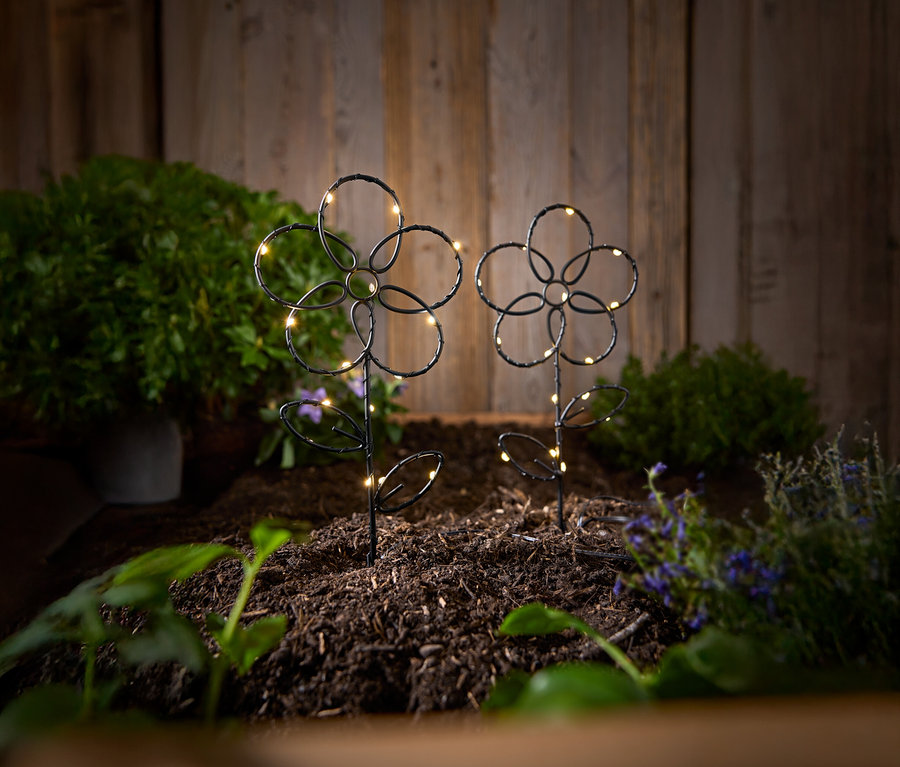 Két fekete, virág alakú kerti karó LED-es lámpákkal áll a földben egy fapalánk előtt.