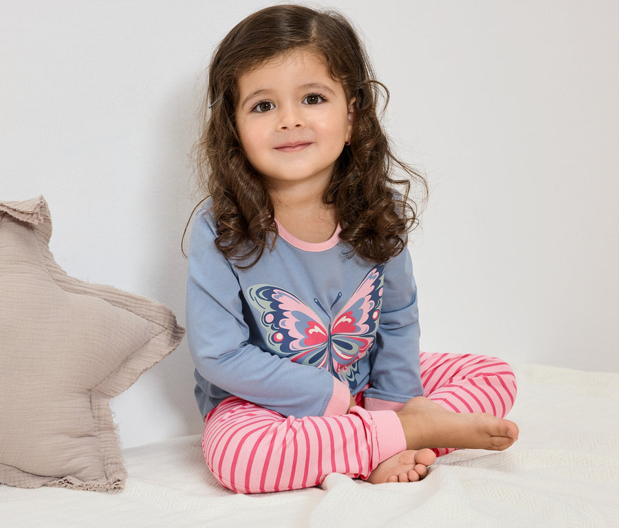 Egy göndör hajú kislány ül az ágyon, kék pillangós pizsamában és rózsaszín csíkos nadrágban.